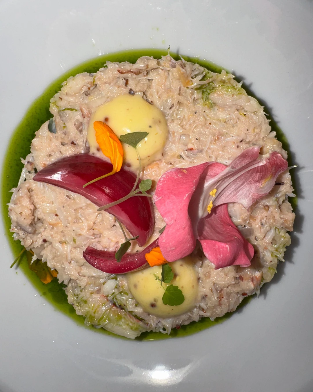 Entrée dressée dans une assiette creuse blanche : disque de préparation, points de crème, pétales roses et orange, huile verte