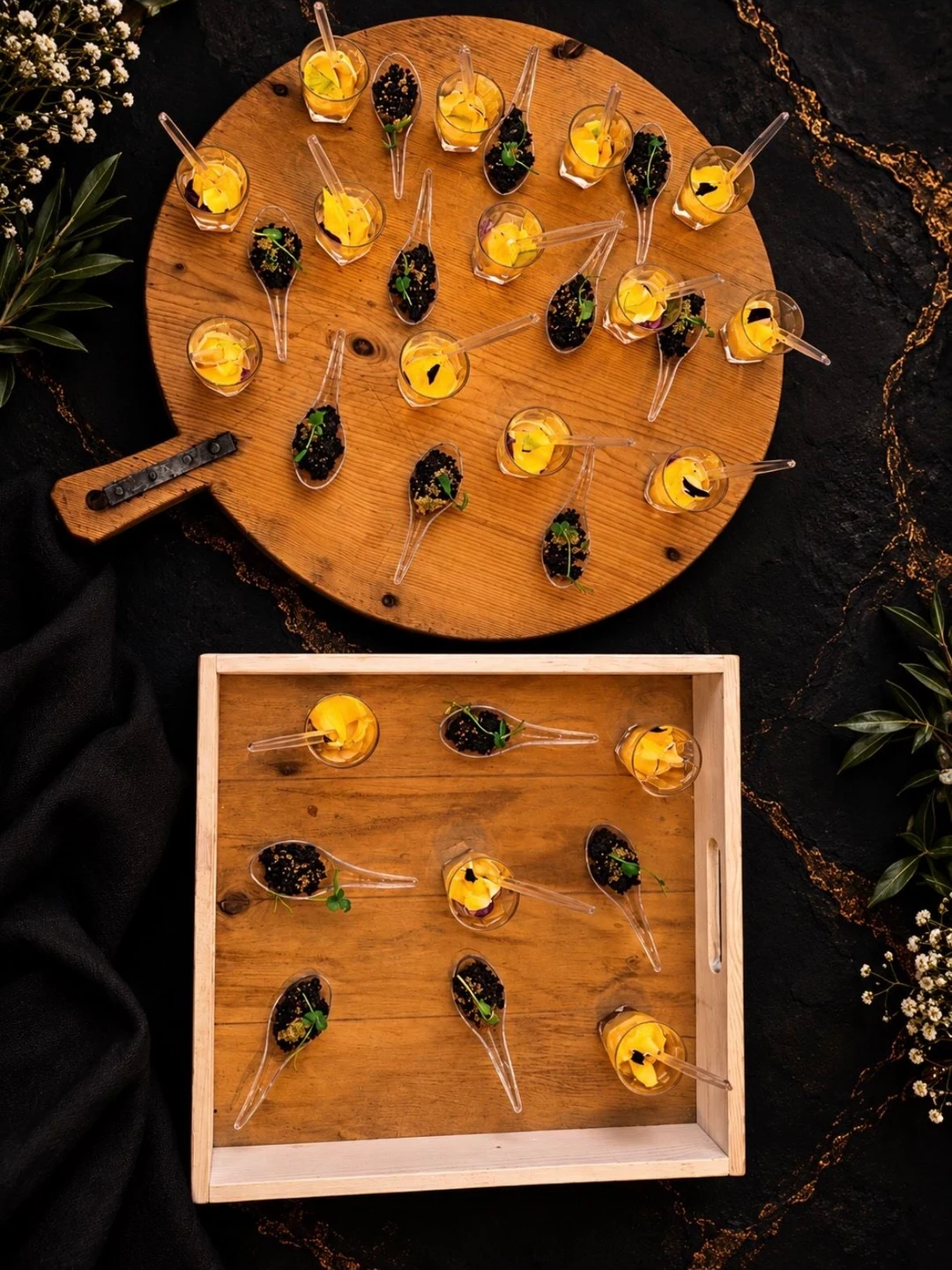 Amuse-bouches en verrines et en cuillères sur une planche ronde et un plateau en bois, fond de marbre noir veiné d’or